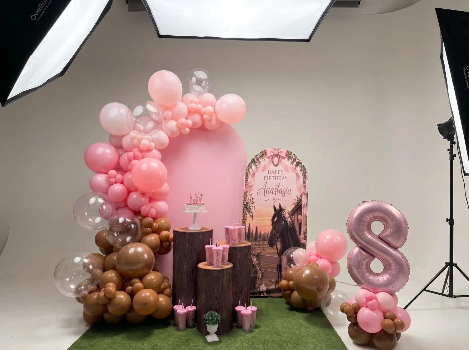 Arco rosa con globos marrones y transparentes, troncos con vasos y una cifra 8 para un cumpleaños con temática de caballos